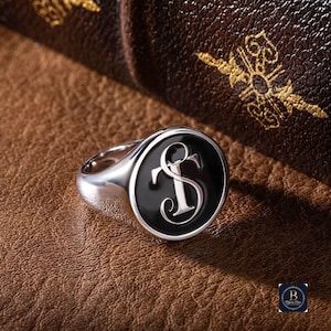 Puede incluir: Un anillo de sello plateado con una cara negra que presenta las letras "S" y "T" en una fuente estilizada. El anillo está sobre una superficie de cuero marrón, con un fondo borroso de un libro encuadernado en cuero.