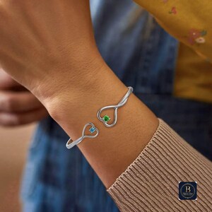 May include: A silver cuff bracelet with heart-shaped terminals, each holding a gemstone. One heart features a blue gem, the other a green gem. The bracelet is designed to be worn on the wrist as jewellery.