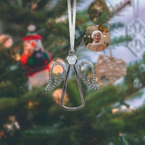 May include: Silver angel Christmas ornament with crystal-studded wings and a white gemstone head. A small photo of a woman is visible in the background. The ornament hangs from a white ribbon against a blurred Christmas tree.