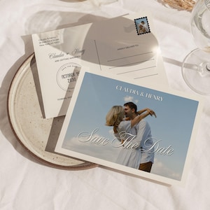 May include: A Save the Date postcard featuring a photograph of a couple embracing against a blue sky. The text reads Claudia & Henry and Save the Date. The reverse side includes address lines, a stamp graphic, and the date October 15, 2030.