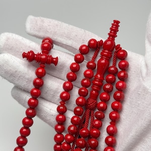 Könnte beinhalten: Ein Set aus leuchtend roten Gebetsperlen mit glatten, kugelförmigen Perlen. Die Schnur enthält ein zentrales, längliches Dekorationselement und eine Quaste aus passender roter Kordel, mit einem markanten kreuzförmigen Teil am Ende.