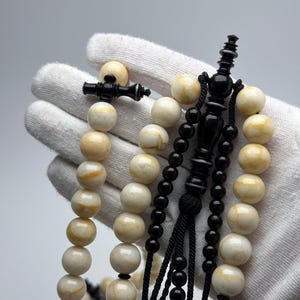 Pode incluir: Um terço com contas redondas e lisas de cor creme com veios amarelos, intercaladas com contas pretas mais pequenas e polidas. Inclui um separador ornamental preto central e uma borla preta com pequenas contas pretas.
