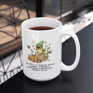 May include: White ceramic mug featuring a whimsical illustration of a frog wearing a rainbow hat on a bed, surrounded by books. The text reads: "MY BED IS A MAGICAL PLACE WHERE I SUDDENLY REMEMBER EVERYTHING I FORGOT TO DO."
