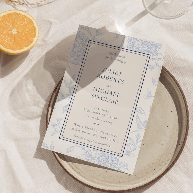 May include: A wedding invitation with a floral design in light blue and white. The invitation includes the names Juliet Roberts and Michael Sinclair, along with the date and time of the wedding. The invitation is on a plate with a lemon slice.