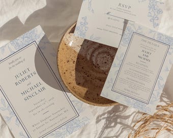 May include: A wedding invitation suite featuring a main invitation, an RSVP card, and a details card, all printed on white cardstock with a light blue floral border and dark blue text. The invitation lists Juliet Roberts and Michael Sinclair.