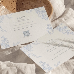 Puede incluir: Dos tarjetas RSVP con diseños florales en las esquinas en azul claro. Las tarjetas tienen campos para la información de los invitados y un código QR. Están sobre una superficie texturizada blanquecina.