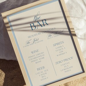 Pode incluir: Uma placa de menu de bar emoldurada com fundo bege claro e texto azul escuro. A placa lista categorias como Signature Cocktails, Wine, Beer, Spirits e Zero Proof, com nomes de bebidas específicos como The Juliet, The Michael e várias bebidas.