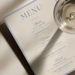 Könnte beinhalten: Eine rechteckige Menükarte mit hellblauem Rand auf cremefarbenem Papier, mit dunkelblauem Text, der Starters, Entrees und Dessert auflistet. Das Menü enthält Gerichte wie Tuna Tartare, Lobster Bisque, Filet Mignon und Wedding Cake, platziert neben einem Glas.