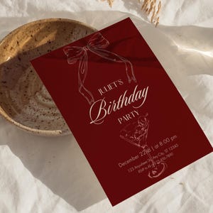 Può includere: Un invito di compleanno bordeaux con scritte e illustrazioni bianche. L'invito recita "Festa di compleanno di Juliet" con i dettagli dell'evento. Un fiocco e un bicchiere da cocktail sono presenti nel design.