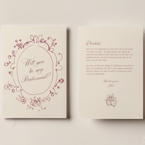 May include: A cream-colored folded card featuring a dark red hand-drawn floral frame and the text Will you be my Bridesmaid? The interior displays a message to Elizabeth, signed by Juliet, with a small illustration of two heart-shaped lockets.
