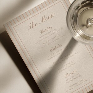 May include: A rectangular menu card with a cream-colored textured surface and thin vertical peach-colored stripes along the edges. The text lists The Menu, Starters, Entrees, and Dessert with specific food items printed in a dark brown serif font.