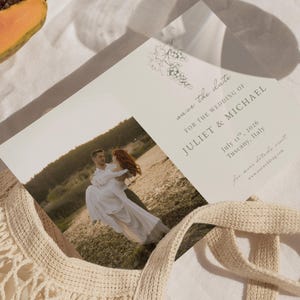May include: Wedding invitation featuring a photo of a couple embracing in a field. The invitation reads "Save the date" and includes the names Juliet & Michael, the date July 11th, 2026, and location Tuscany, Italy. A floral illustration is at the top.