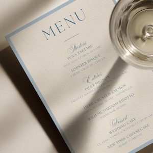 May include: A rectangular menu card with a light blue border on cream-colored textured paper. The text is printed in blue ink, listing sections for Starters, Entrees, and Dessert, including items like Tuna Tartare, Filet Mignon, and Wedding Cake.