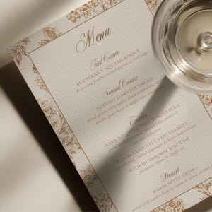 Könnte beinhalten: Eine rechteckige Menükarte aus Papier mit einem kupferfarbenen Blumenrand. Der Text listet Gänge wie Butternusskürbissuppe, Herbstsalat, Kräuter-Rinderfilet, gebratenen Atlantiklachs, Wildpilzrisotto und warmen Apfel-Crumble auf.