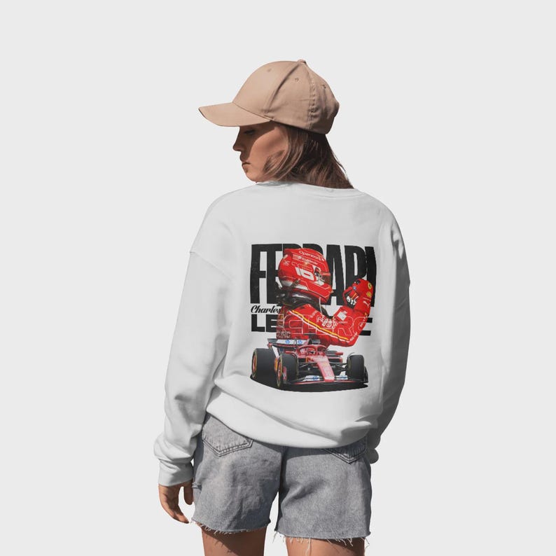 Puede incluir: Sudadera blanca con un gr&aacute;fico de un coche de carreras Ferrari rojo y su piloto. La palabra "FERRARI" est&aacute; impresa en letras negras grandes encima de la imagen. El piloto lleva un casco y un traje de carreras rojos.