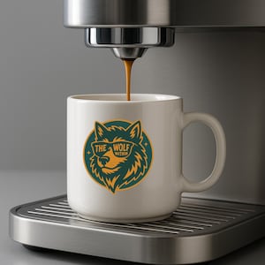 Puede incluir: Una taza de cerámica blanca con asa, con un diseño gráfico circular de la cabeza de un lobo con gafas de sol, con el texto "THE WOLF WITHIN". La taza está debajo de una cafetera.