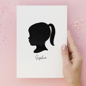 May include: A white notebook featuring a black silhouette of a child's profile with a ponytail. The name "Sophia" is written in cursive below the silhouette. The notebook is held by a hand against a pink background.