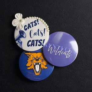 Può includere: Tre spille rotonde. Una spilla bianca presenta il testo "CATS! Cats! CATS!" in blu, un megafono e decorazioni con perle e fiori bianchi. Una spilla blu mostra un gatto selvatico arancione. Una spilla viola riporta la scritta "Wildcats".