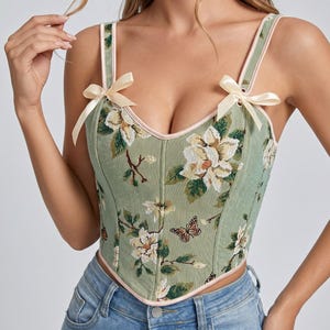 May include: A green floral corset top with cream-colored bows on the straps. The top features a botanical pattern with white flowers and butterflies. The edges are trimmed with a light pink piping.