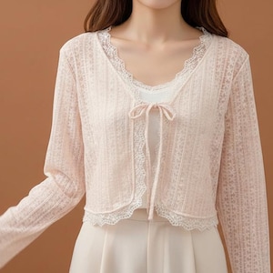 May include: A light pink lace cardigan with long sleeves and a tie closure. The cardigan has a delicate floral pattern and white lace trim along the neckline and hem. The cardigan is worn over a white top.