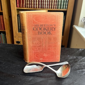 May include: A vintage orange hardback book titled Mrs Beeton's Cookery Book with embossed illustrations of serving dishes on the front cover, accompanied by two silver-toned metal ladles with curved handles resting in a crossed position.