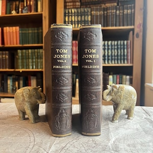 Puede incluir: Dos libros antiguos encuadernados en cuero marrón, "Tom Jones, Vol. 1 y 2, Fielding", se muestran en posición vertical. Las lomos tienen letras doradas y relieves decorativos. Dos pequeñas figuras de elefantes flanquean los libros sobre una superficie impresa con mapas.