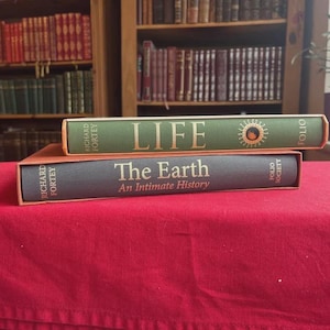 Może przedstawiać: Dwie książki w twardej oprawie, "LIFE" i "The Earth: An Intimate History", ułożone na czerwonej powierzchni. Górna książka jest zielona ze złotymi literami i ilustracją słońca. Dolna książka jest szara ze złotymi literami.