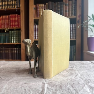 May include: A vintage book titled "Saint Jude" with a light yellow cover, propped up by a brass camel figurine. The book is positioned on a map-printed surface, with bookshelves in the background.