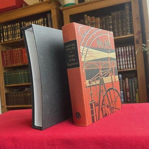 May include: Two hardcover books on a red surface. The book on the right has an orange cover with the title "Lives of the Engineers" by Samuel Smiles. The cover features a red and black illustration of machinery. The book on the left has a black cover.