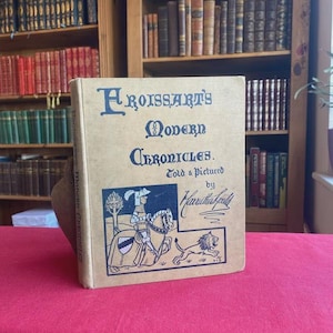 May include: A vintage book titled "Froissart's Modern Chronicles" with blue lettering. The cover features a black and white illustration of a knight on horseback and a lion. The book is propped up on a red surface, with bookshelves in the background.