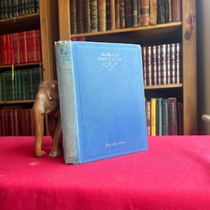 May include: A light blue hardcover book titled The Works of Jane Austen, specifically the volume Persuasion, featuring embossed silver lettering on the front cover and spine, positioned next to a small carved wooden elephant figurine.