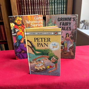 May include: Three vintage hardcover books titled Peter Pan, Girls Adventure Stories, and Grimm's Fairy Tales. The front book features Peter Pan flying over a ship, while the two books behind display illustrated scenes with characters and text.