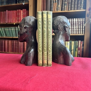 Puede incluir: Un par de sujetalibros de madera marrón oscuro, cada uno esculpido con la forma de una cabeza humana de perfil. Tres libros antiguos con cubiertas verdes y letras doradas se colocan entre los sujetalibros. Los libros se titulan "Amelia" y son de la editorial J.M. Dent & Sons.