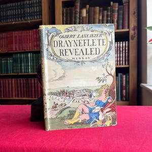 Pode incluir: Um livro vintage intitulado "Drayneflete Revealed" de Osbert Lancaster. A capa apresenta uma ilustração colorida de uma mulher em um vestido azul e botas amarelas, reclinada perto de uma árvore com uma vista da cidade ao fundo. O livro está em uma superfície vermelha.