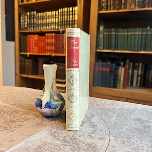 Op de afbeelding: Een vintage boek met de titel "Allan Quatermain" met een rode rug en lichtgroene kaft staat rechtop naast een decoratieve vaas met blauwe en roze bloemmotieven. Het boek heeft gouden accenten. Boekenkasten vol boeken staan op de achtergrond.