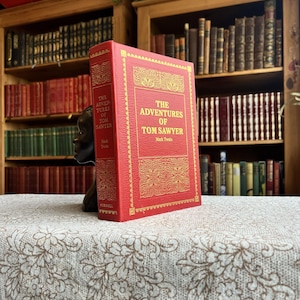 Op de afbeelding: Een rood hardcover boek met de titel "The Adventures of Tom Sawyer" met gouden letters, ondersteund door een zwarte buste boekensteun. Het boek ligt op een tafelkleed met bloemenpatroon, met boekenplanken op de achtergrond.