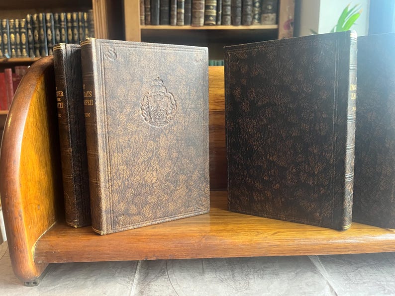 May include: A collection of antique books with textured brown covers, displayed on a wooden shelf. The books have embossed designs and are arranged in a wooden book holder. The background features a bookshelf filled with more books.