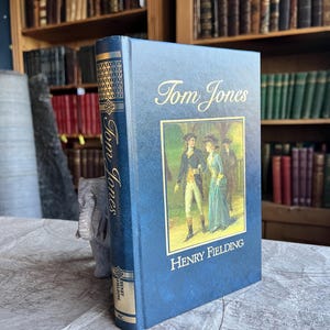Puede incluir: Un libro de tapa dura azul titulado "Tom Jones" de Henry Fielding. El libro tiene letras doradas y una ilustración a color. El lomo muestra el título y el autor. El libro está sobre un mapa.