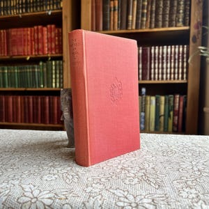 May include: A coral-coloured hardcover book titled "The Sayard of India" by Potter. The book is standing upright on a floral patterned tablecloth. Bookshelves filled with books are in the background.