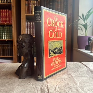 Op de afbeelding: Een hardcover boek getiteld "The Crock of Gold" van James Stephens, met een rode kaft en gouden letters. Het boek wordt ondersteund door een donkere houten sculptuur. De achtergrond toont een boekenkast vol boeken.