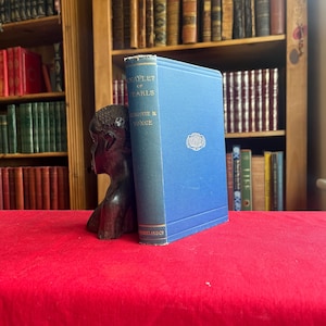 Op de afbeelding: Een blauw hardcover boek getiteld Chaplet of Pearls door Charlotte M. Yonge met gouden letters op de rug, geplaatst naast een donkerbruine gesneden houten buste van een persoon. Het boek heeft een klein decoratief embleem op de voorkant.