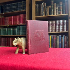 May include: A maroon-colored book with embossed text and a carved wooden elephant figurine on a red tablecloth. The book is propped up against the elephant. Bookshelves filled with books are in the background.