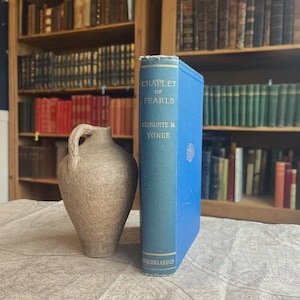 Op de afbeelding: Een stilleven met een blauw boek getiteld "Chaplet of Pearls" van Charlotte M. Yonge, naast een beige keramische kruik. Het boek staat rechtop op een oppervlak met kaartpatroon, met boekenplanken op de achtergrond.
