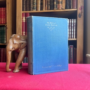 May include: A blue hardcover book titled The Works of Jane Austen: Sense and Sensibility with gold lettering and a small decorative emblem, positioned next to a carved wooden elephant figurine on a flat red surface.