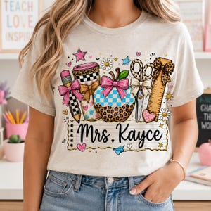 A woman wearing a white t-shirt with a colorful graphic design featuring various school supplies and the name "Mrs. Kayce".