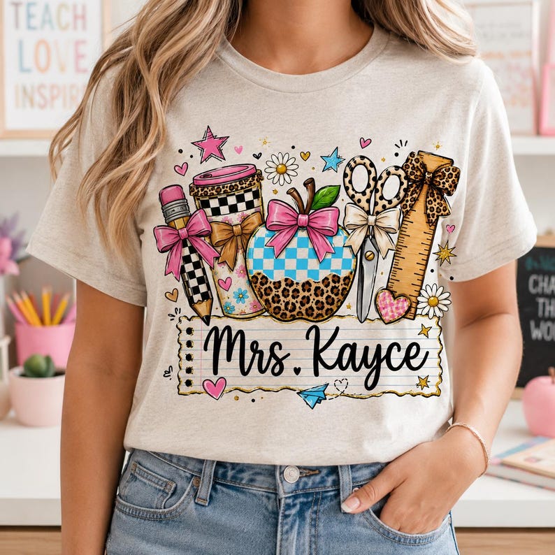 A woman wearing a white t-shirt with a colorful graphic design featuring various school supplies and the name "Mrs. Kayce".
