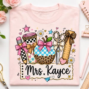 A pink t-shirt with a colorful illustration of various school supplies and a personalized name, "Mrs. Kayce", printed on it.
