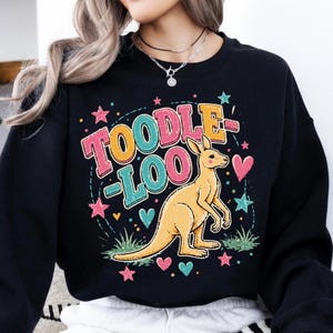 Op de afbeelding: Zwart sweatshirt met een kleurrijke afbeelding. Het ontwerp toont de woorden "TOODLE LOO" in roze, geel en teal, met een kangoeroe-illustratie. Roze sterren en harten omringen de tekst en de kangoeroe.