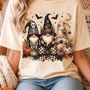 May include: A cream-colored short-sleeve t-shirt featuring a graphic of three gnomes dressed for Halloween. One gnome wears a skeleton costume, one a witch hat, and one is wrapped like a mummy. The design includes pumpkins, bats, and a bow.