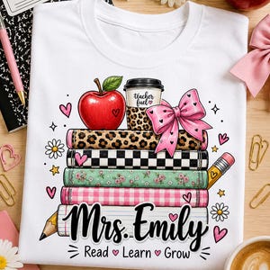 Könnte beinhalten: Ein weißes T-Shirt mit einer Grafik aus fünf gestapelten Büchern, einem roten Apfel, einem Kaffeebecher mit der Aufschrift teacher fuel und einer rosa Schleife. Der Text Mrs. Emily steht in schwarzer Schrift, darunter Read, Learn, Grow.